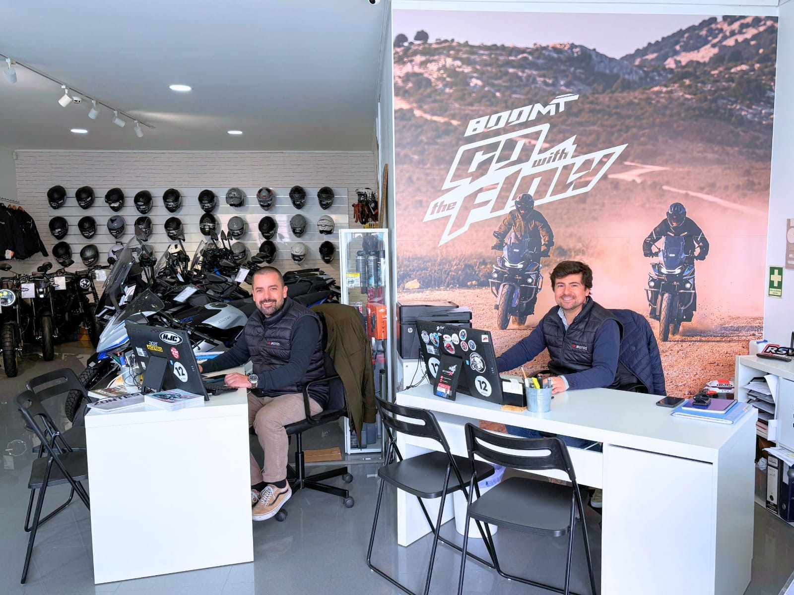 Interior do concessionário da INMOTOS. Vê-se o dono e o parceiro da INMOTOS, sentados atrás de secretárias. Atrás do dono, vê-se uma parede com um poster da moto 800MT. Atrás do parceiro, vêm-se capacetes de motas e motos.